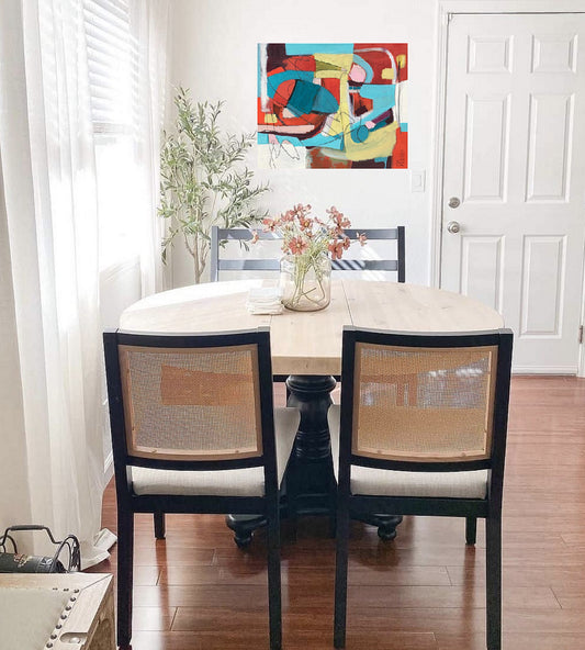 Red, yellow, aqua and white abstract painting on canvas created by artist danielle nelisse installed on white wall in modern dining room in front of contemporary dining set with vase of flowers on table