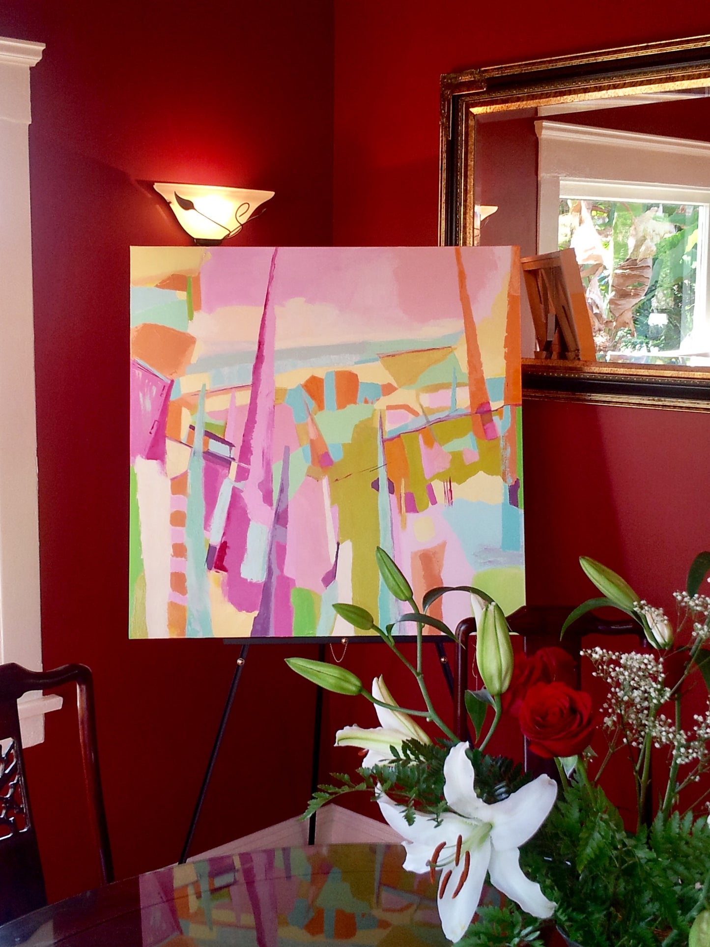 Sage and pink abstract oil painting displayed on easel in dining room with burgundy walls, gold framed mirror, and fresh bouquet of lilies on dining table.