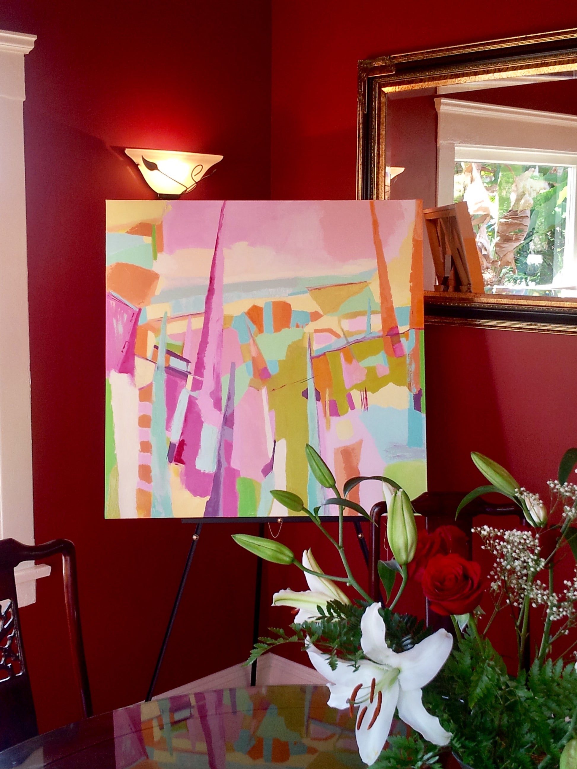 Sage and pink abstract oil painting displayed on easel in dining room with burgundy walls, gold framed mirror, and fresh bouquet of lilies on dining table.